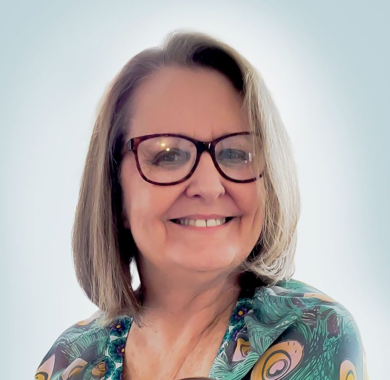 A photograph of Dindy Murphy wearing glasses. Lady wearing peacock featherd blouse.