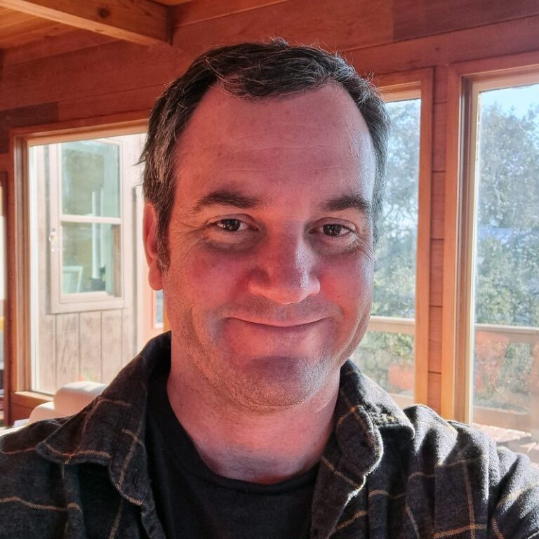 Man smiling wearing a flannel shirt with large windows behind him.